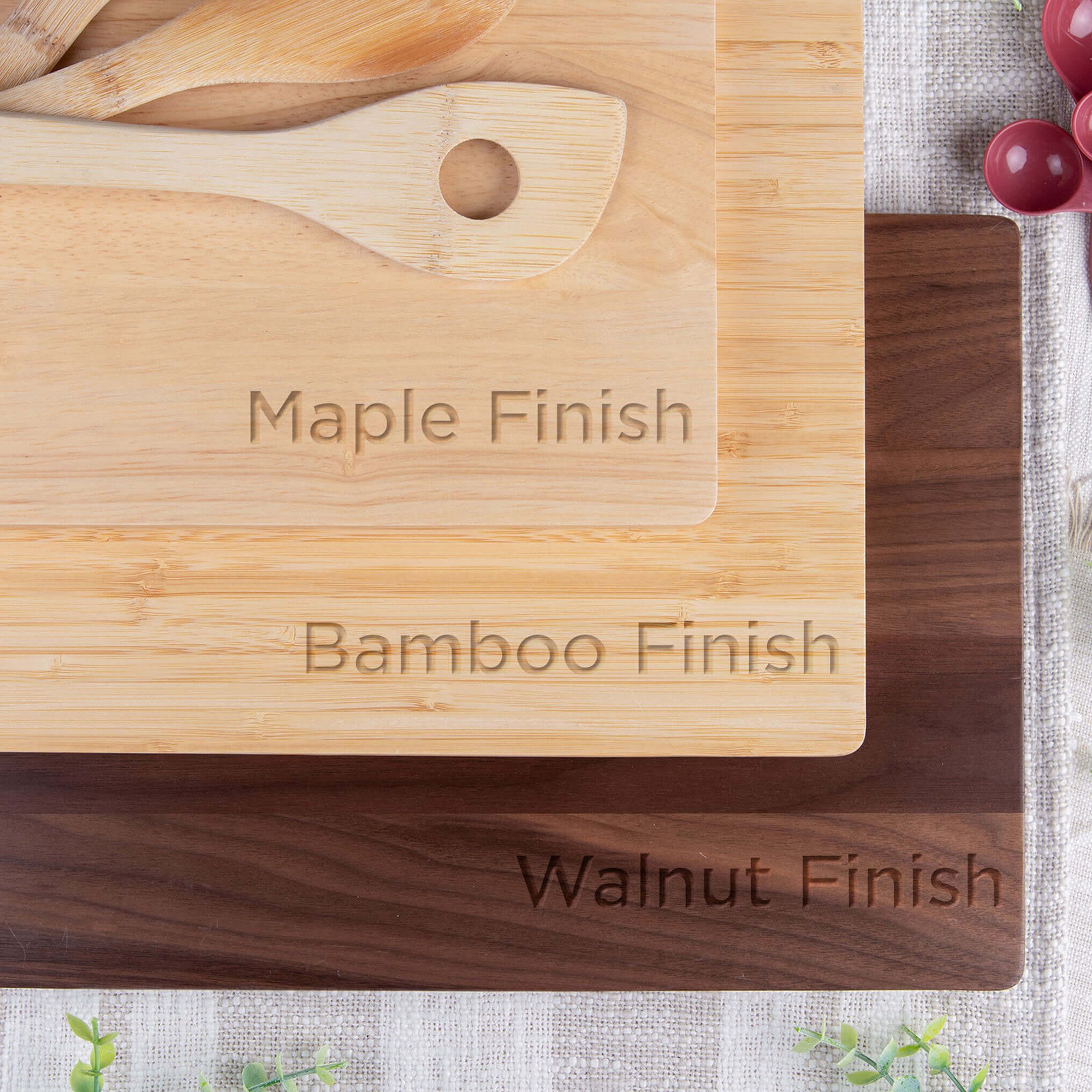 Top-down view of three engraved cutting boards labeled Maple Finish, Bamboo Finish, and Walnut Finish, stacked to show the color and grain differences between each wood type with wooden utensils and red measuring spoons nearby.