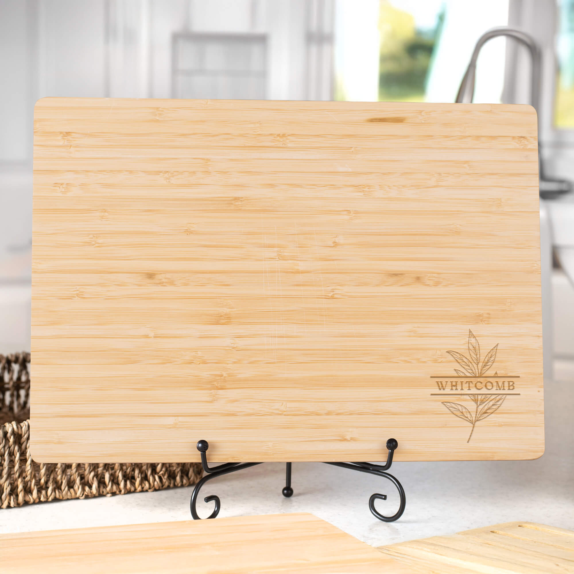 Engraved bamboo cutting board with “Whitcomb” and botanical leaf design, displayed on a black stand in a sunlit kitchen.