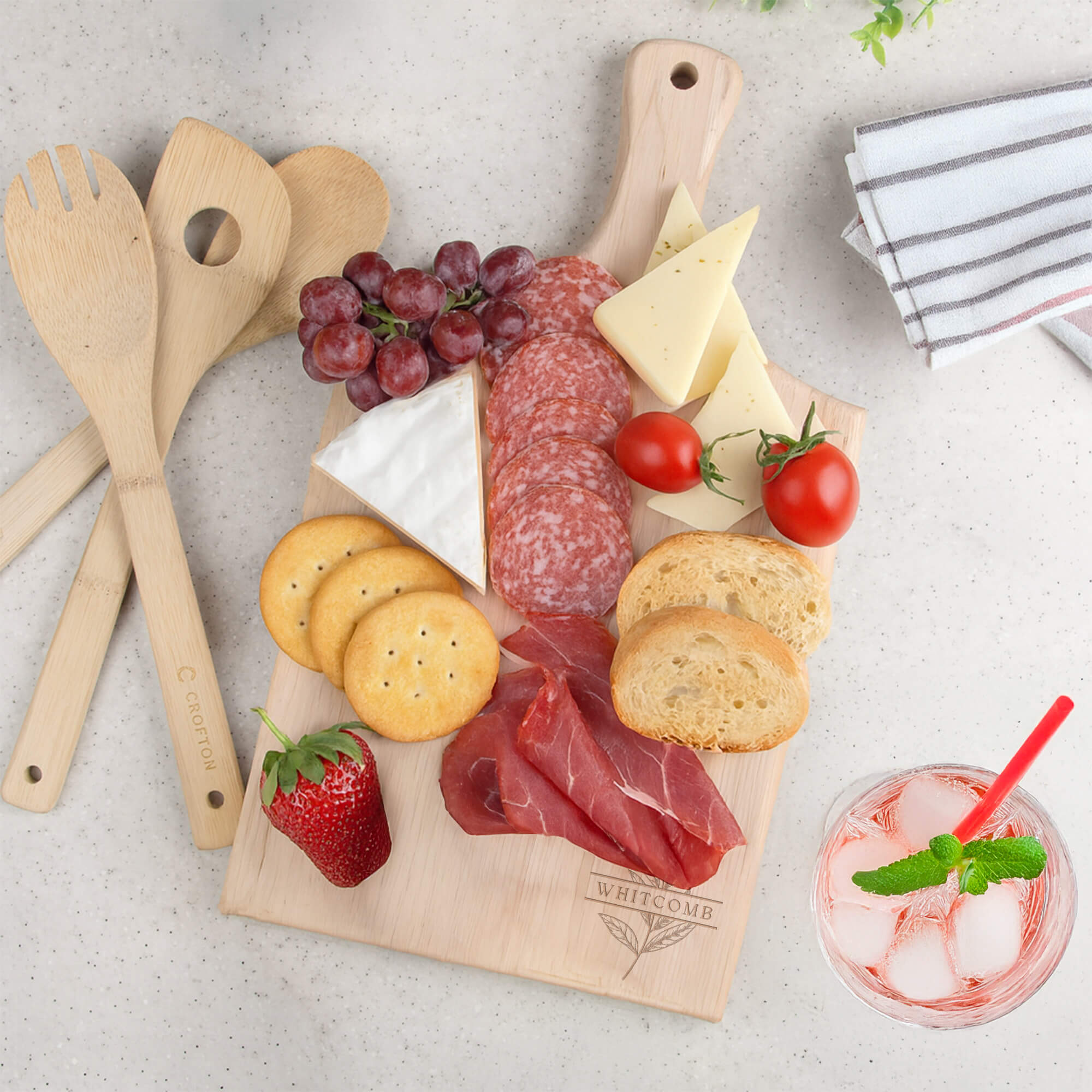 Personalized paddle cutting board engraved with “Whitcomb” and a botanical leaf design, used as a charcuterie board with meats, cheese, and fresh fruit.