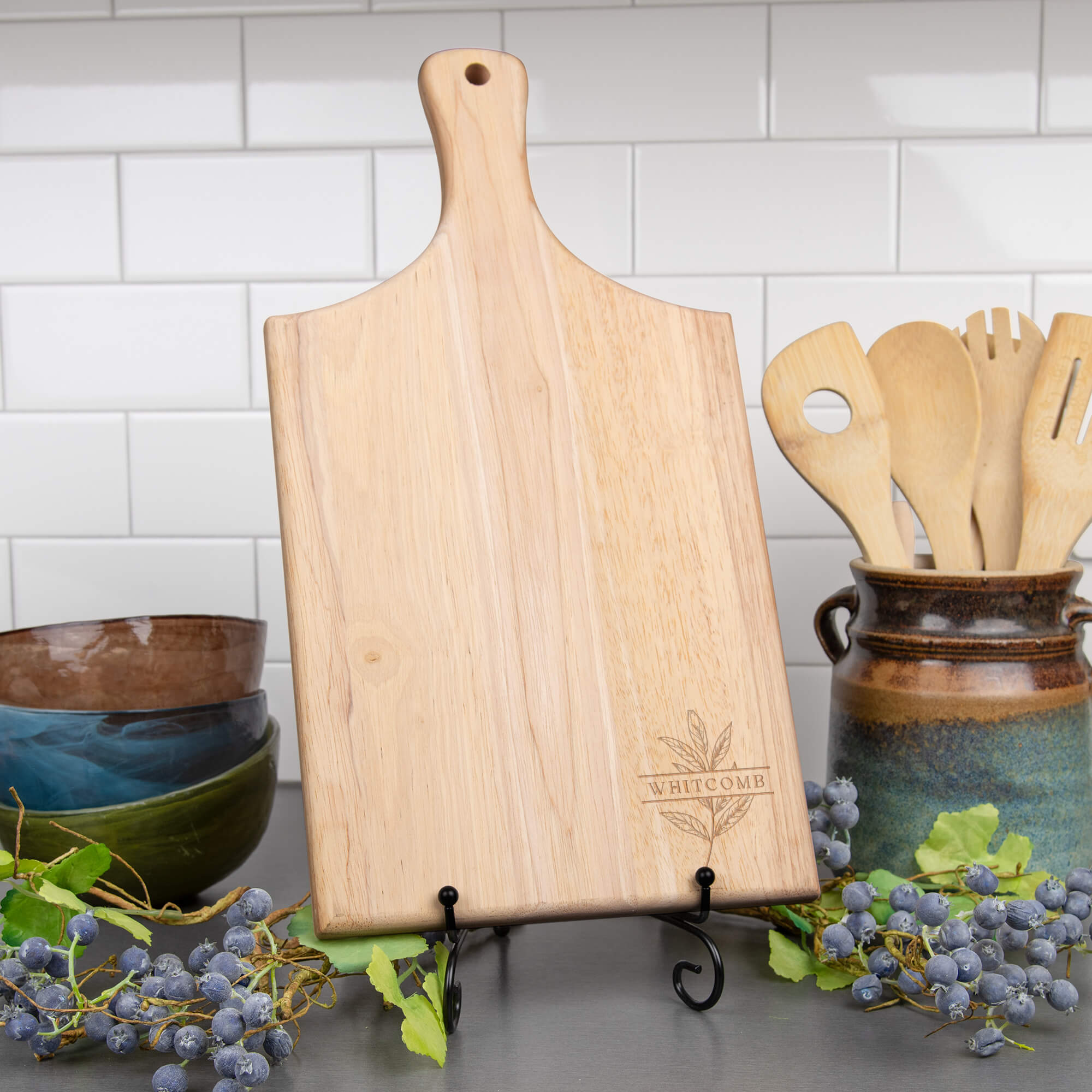 ngraved paddle cutting board featuring “Whitcomb” and a botanical leaf design, displayed upright on a kitchen counter beside ceramic bowls and wooden utensils.
