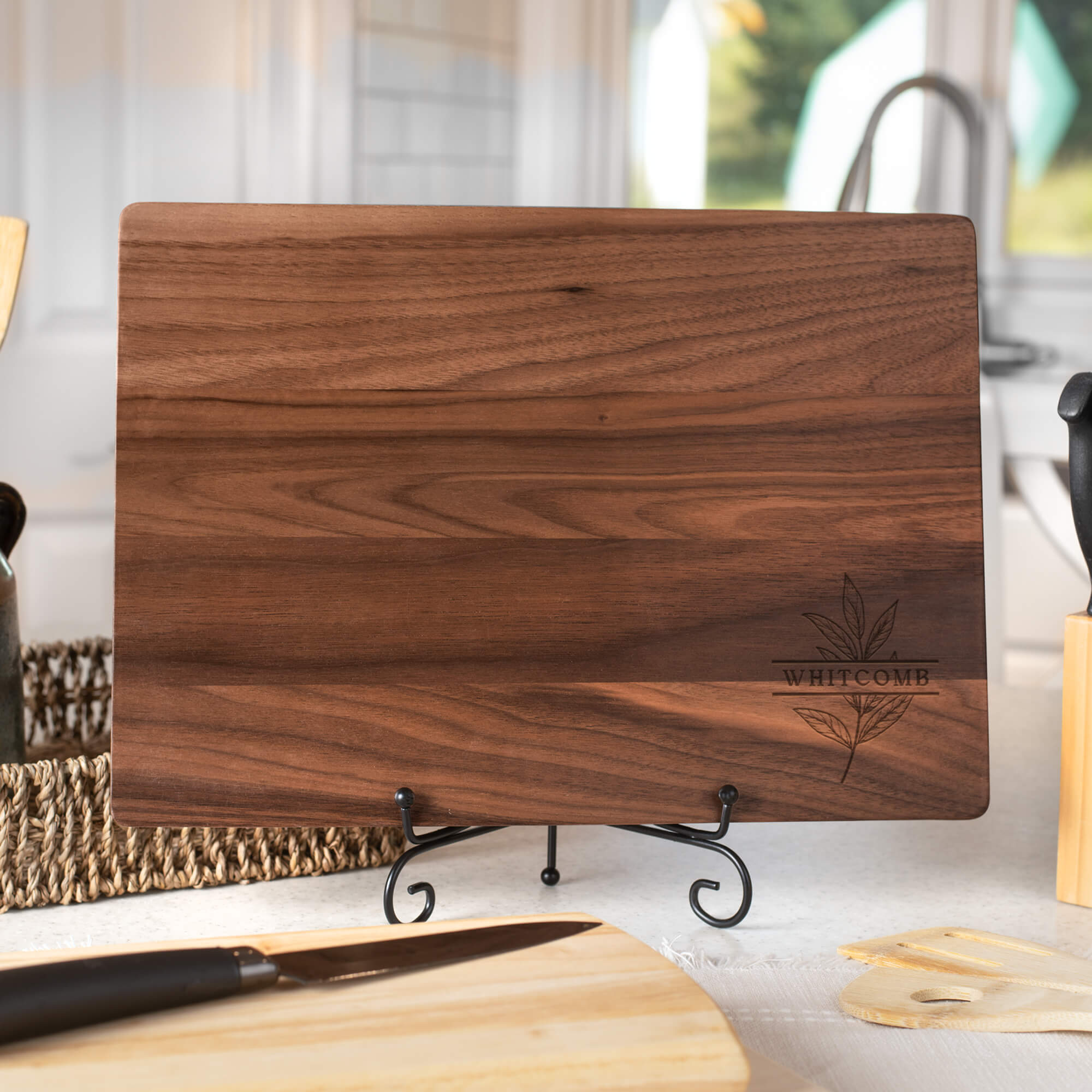 Close-up of personalized walnut cutting board engraved with “Whitcomb” and a botanical leaf design, styled with grapes, cheese, and wine corks.