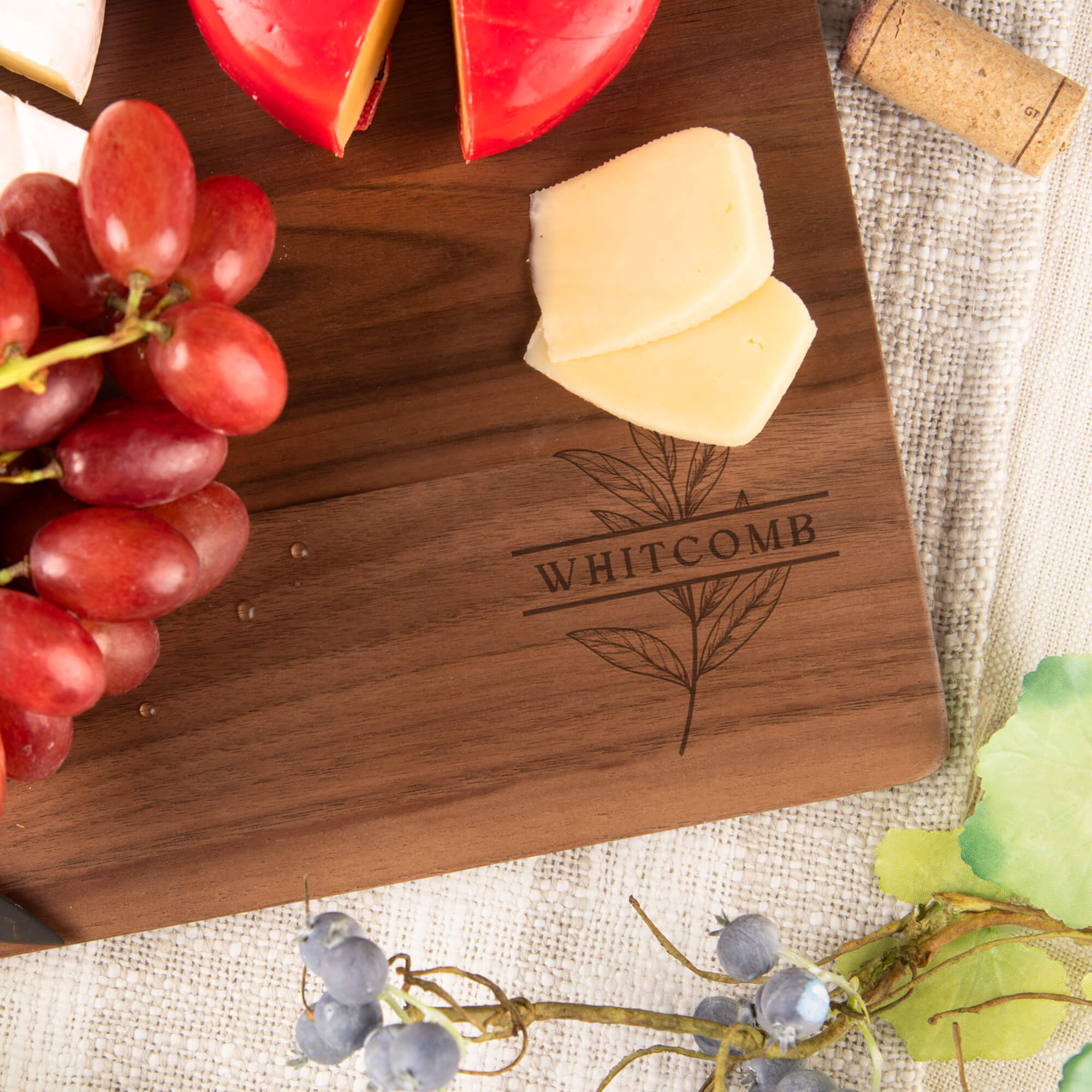 Engraved walnut cutting board featuring “Whitcomb” and a botanical leaf motif, displayed on a kitchen counter with natural light and utensils.