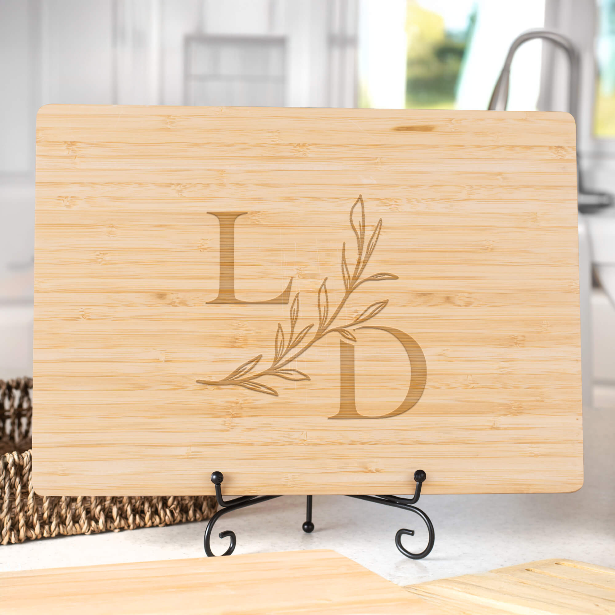 Bamboo cutting board with serif L and D initials linked by a leafy branch on a display stand in a kitchen