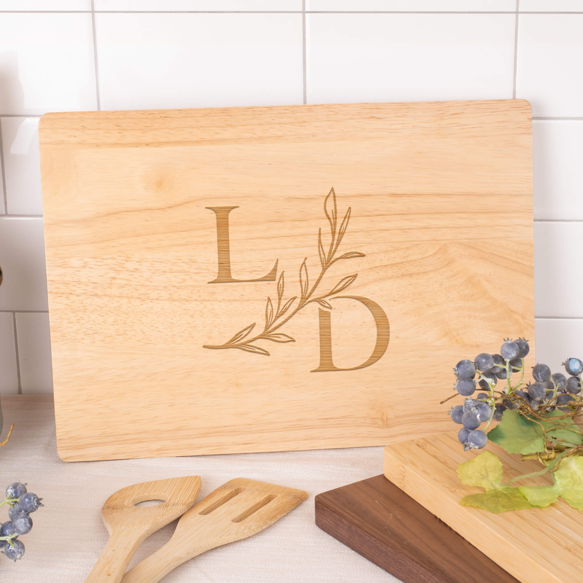 Personalized maple cutting board engraved with serif L and D initials and a leafy accent, styled on a kitchen counter