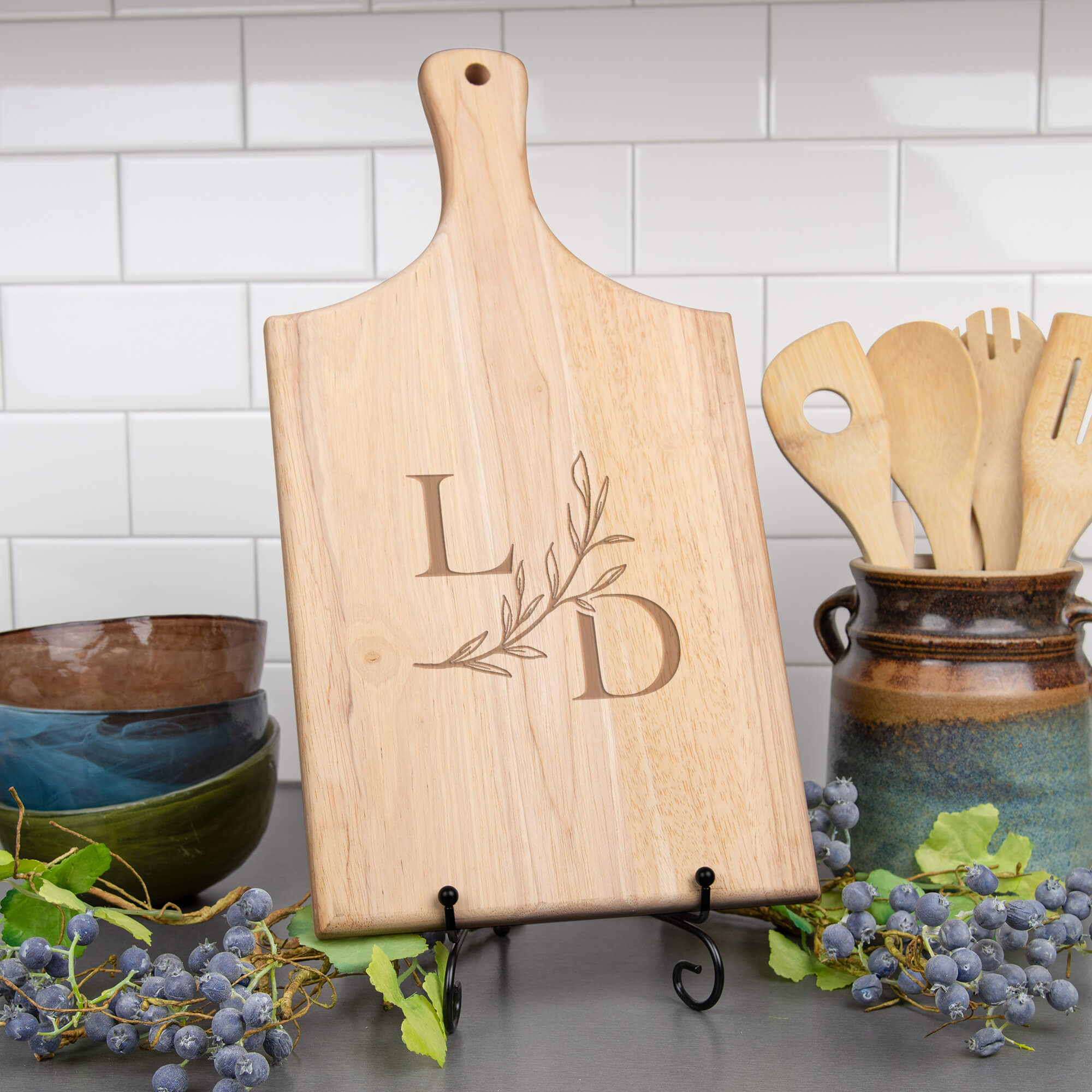 Maple paddle cutting board with serif L and D initials and a sweeping leaf branch displayed on a stand