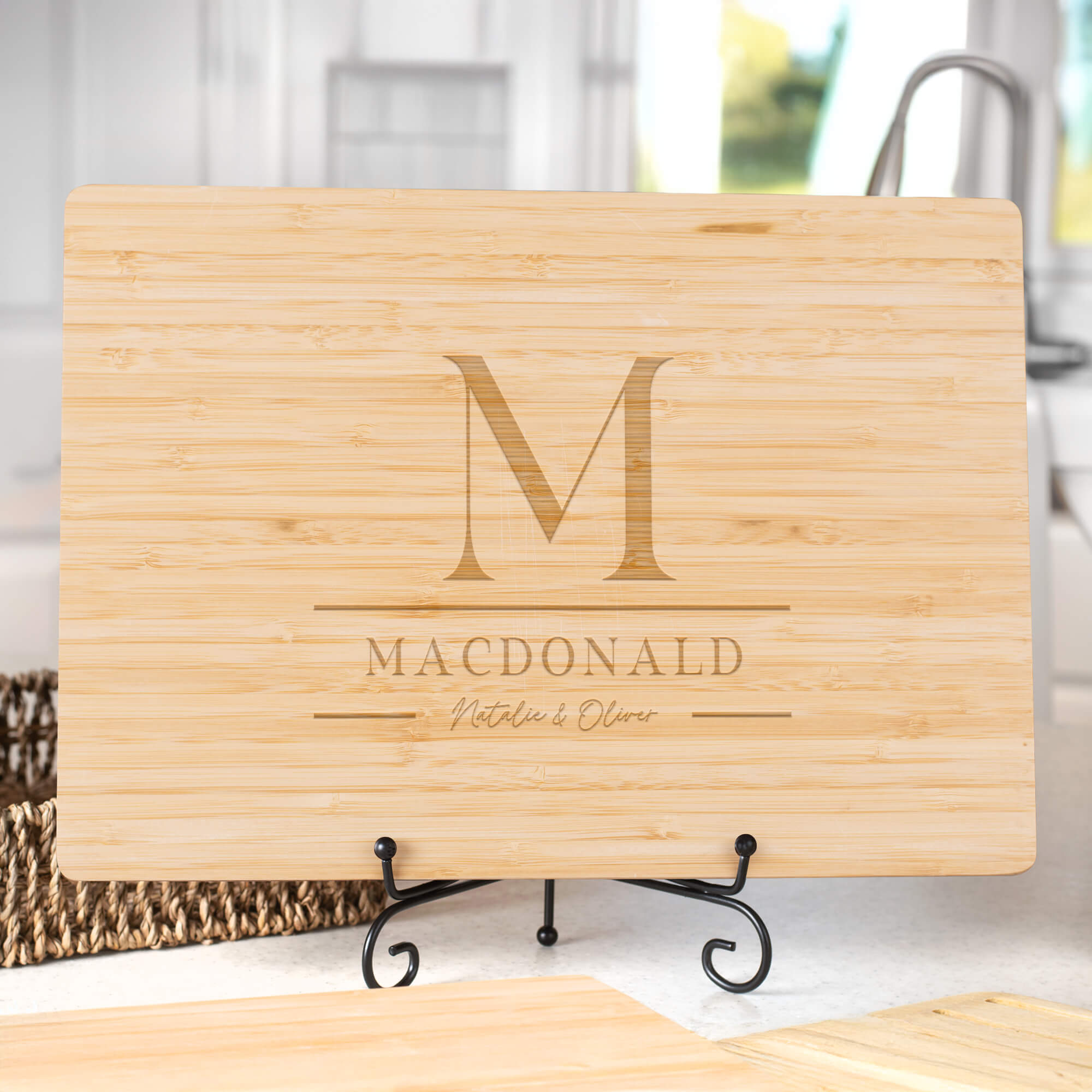 Bamboo cutting board with stacked monogram M, family name MacDonald, and handwritten script names Natalie & Oliver displayed on a stand in a kitchen