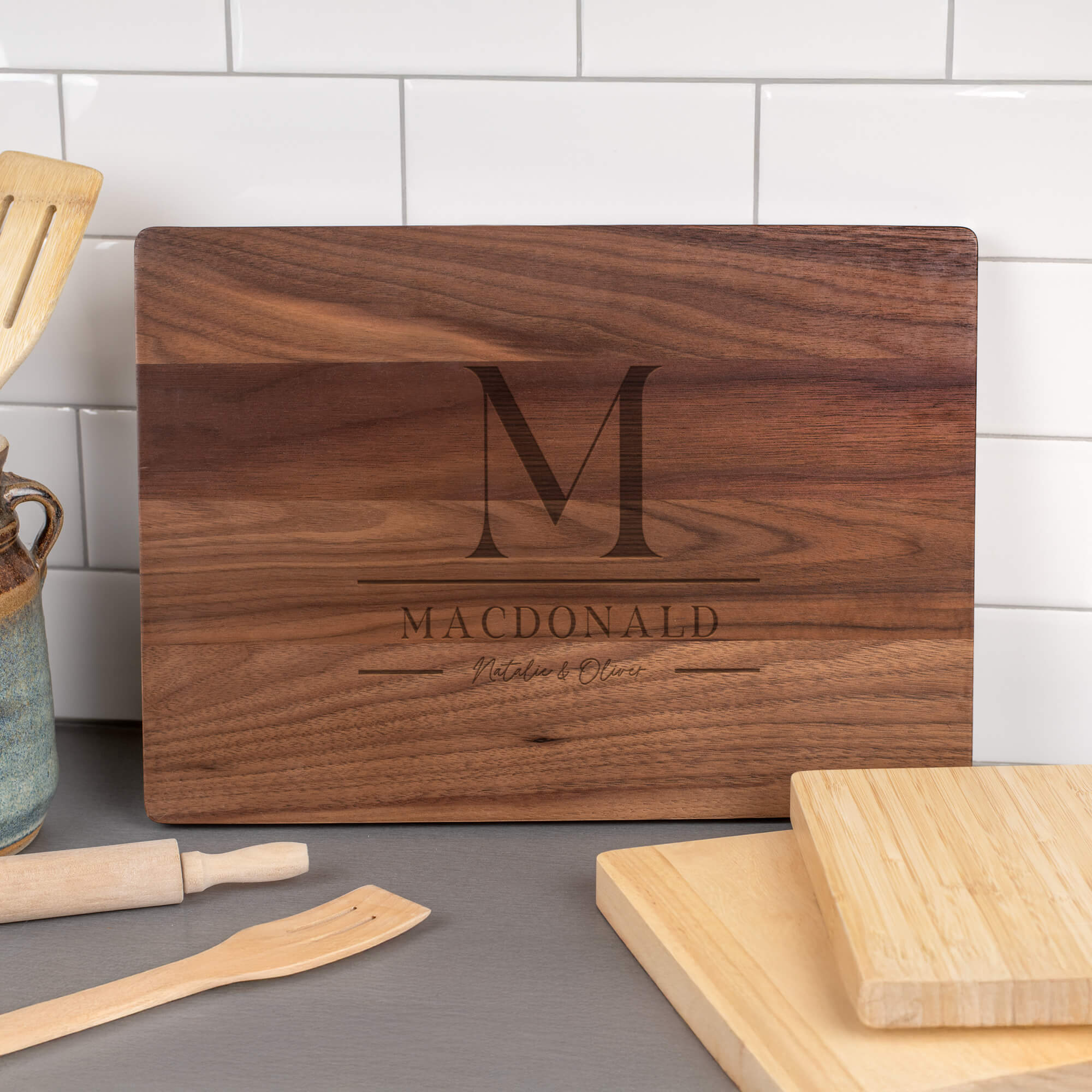 Walnut cutting board engraved with stacked M monogram, MacDonald family name, and script Natalie & Oliver displayed in kitchen setting