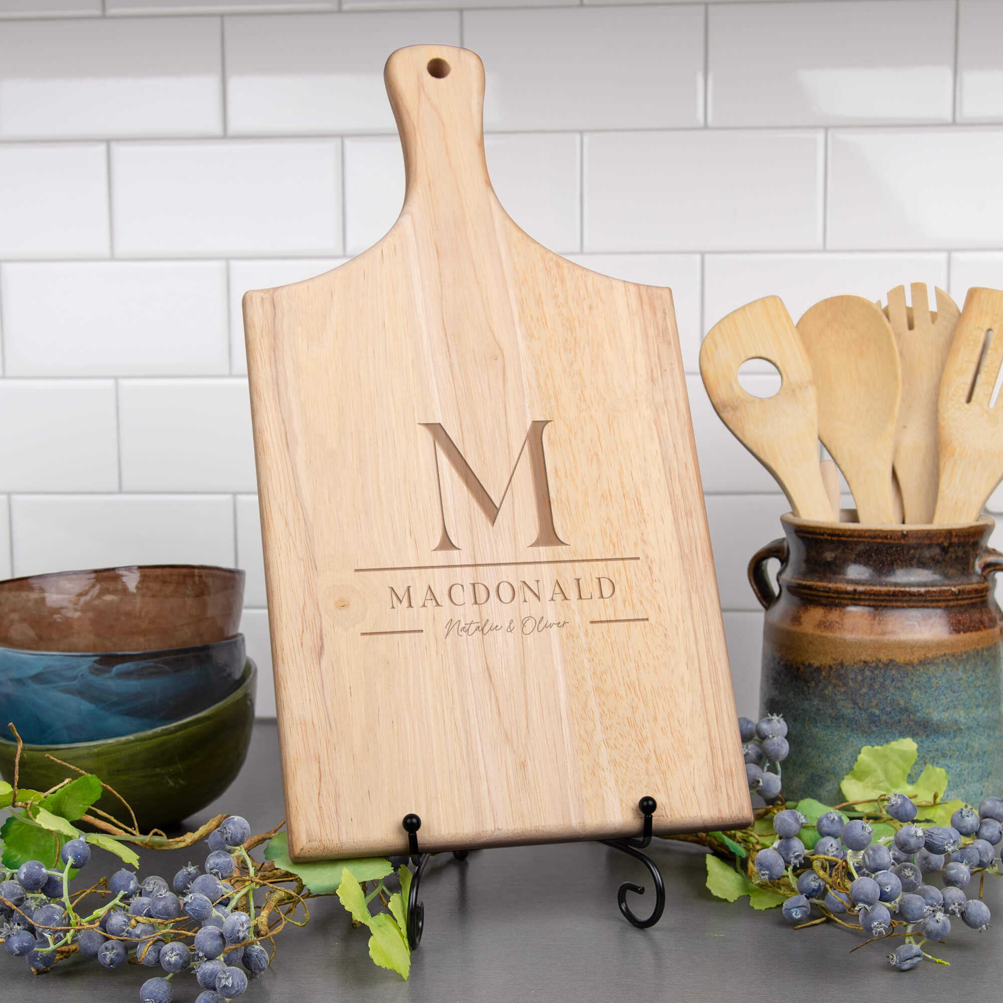 Walnut cutting board with stacked M monogram, MacDonald family name, and Natalie & Oliver in script shown on kitchen counter beside utensils