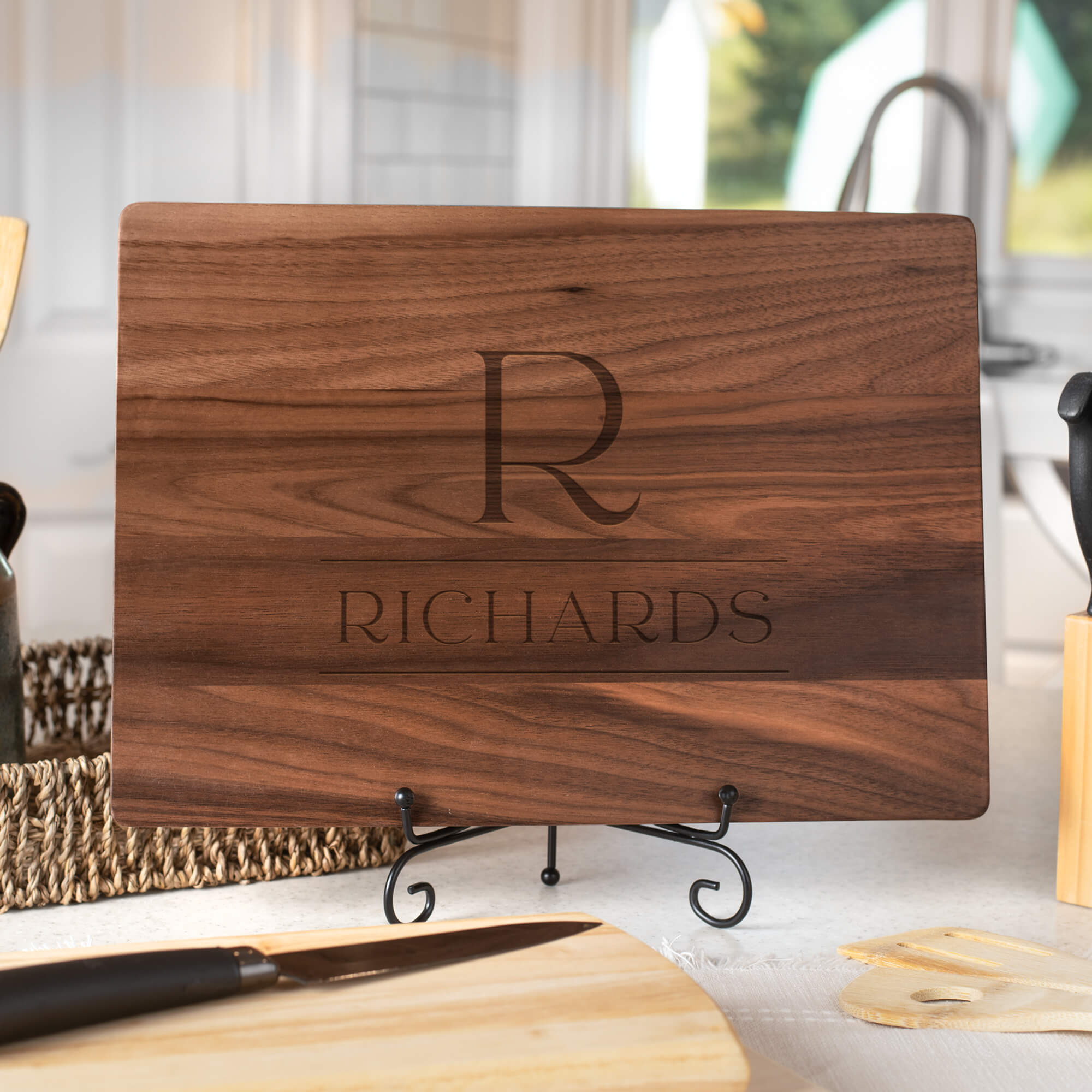 Personalized walnut cutting board with serif R monogram and engraved Richards family name displayed on stand in bright kitchen
