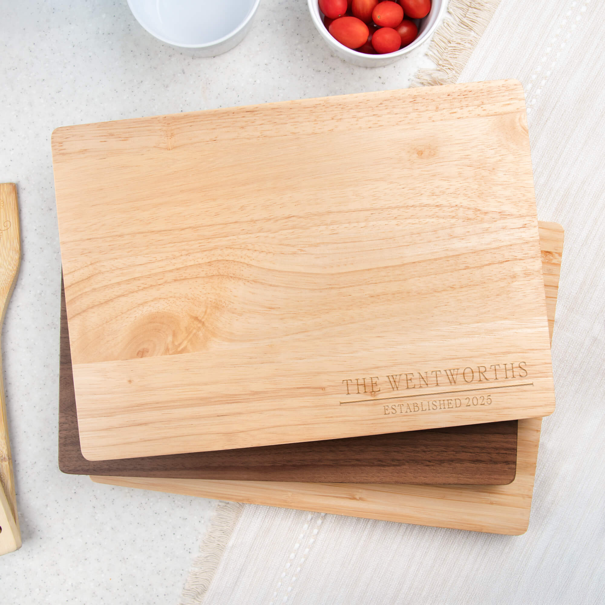 Personalized maple cutting board engraved with “The Wentworths Established 2025” displayed on a kitchen counter with fresh tomatoes and utensils.