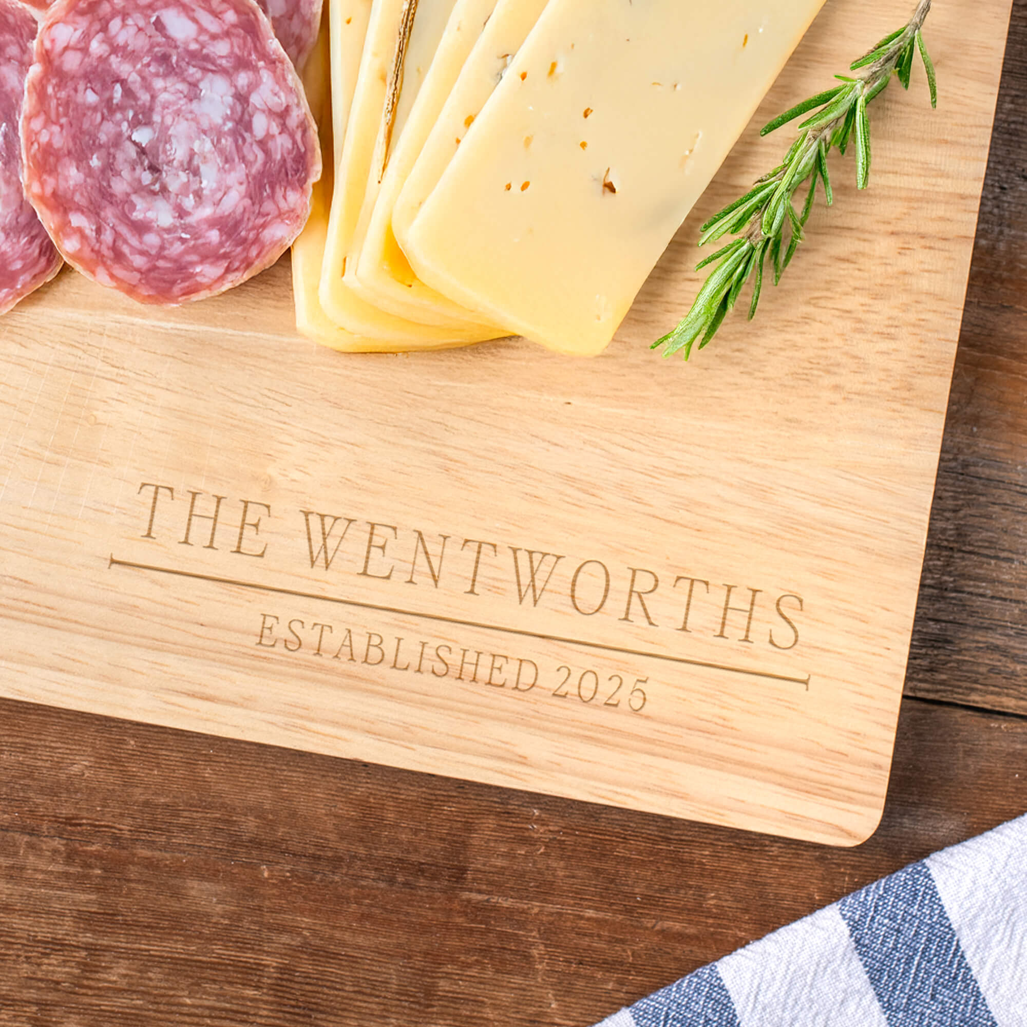 Close-up of custom maple cutting board engraved with “The Wentworths Established 2025,” surrounded by cheese, charcuterie, and rosemary sprigs.