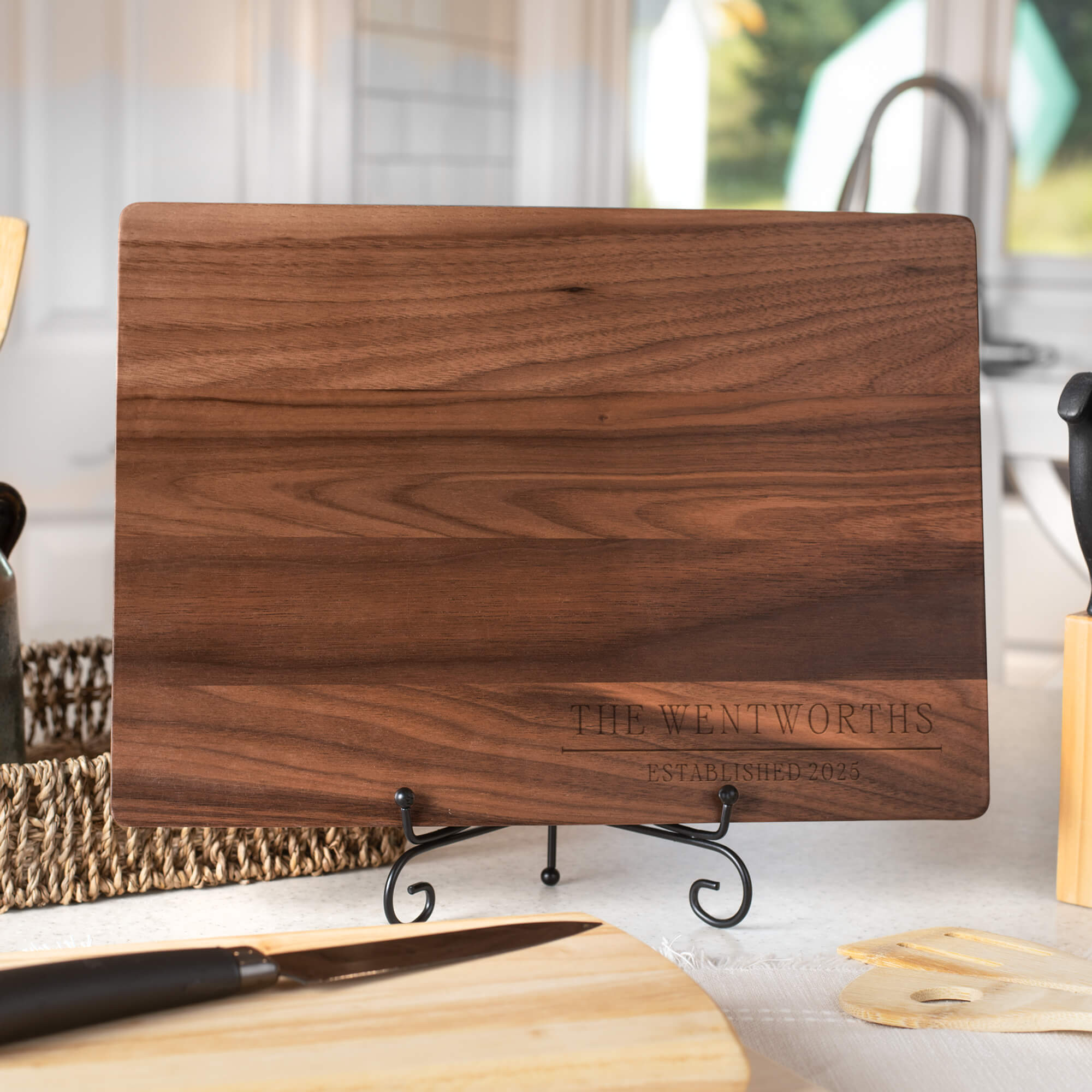 Engraved walnut cutting board featuring “The Wentworths Established 2025,” displayed on a countertop with kitchen utensils and natural light.