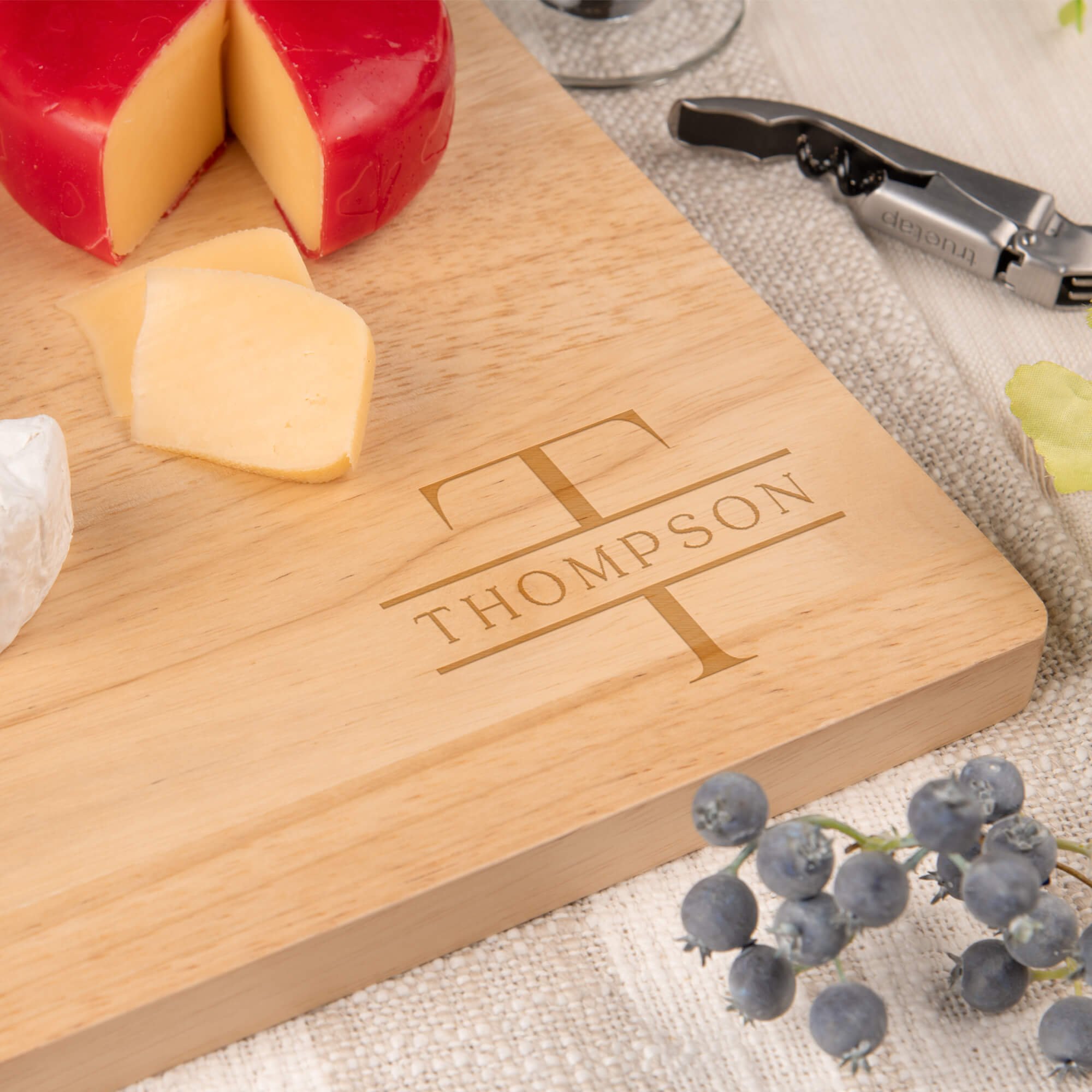 Closeup of corner of maple cutting board with Engragved Family Name Thompson next to an assortment of cheeses