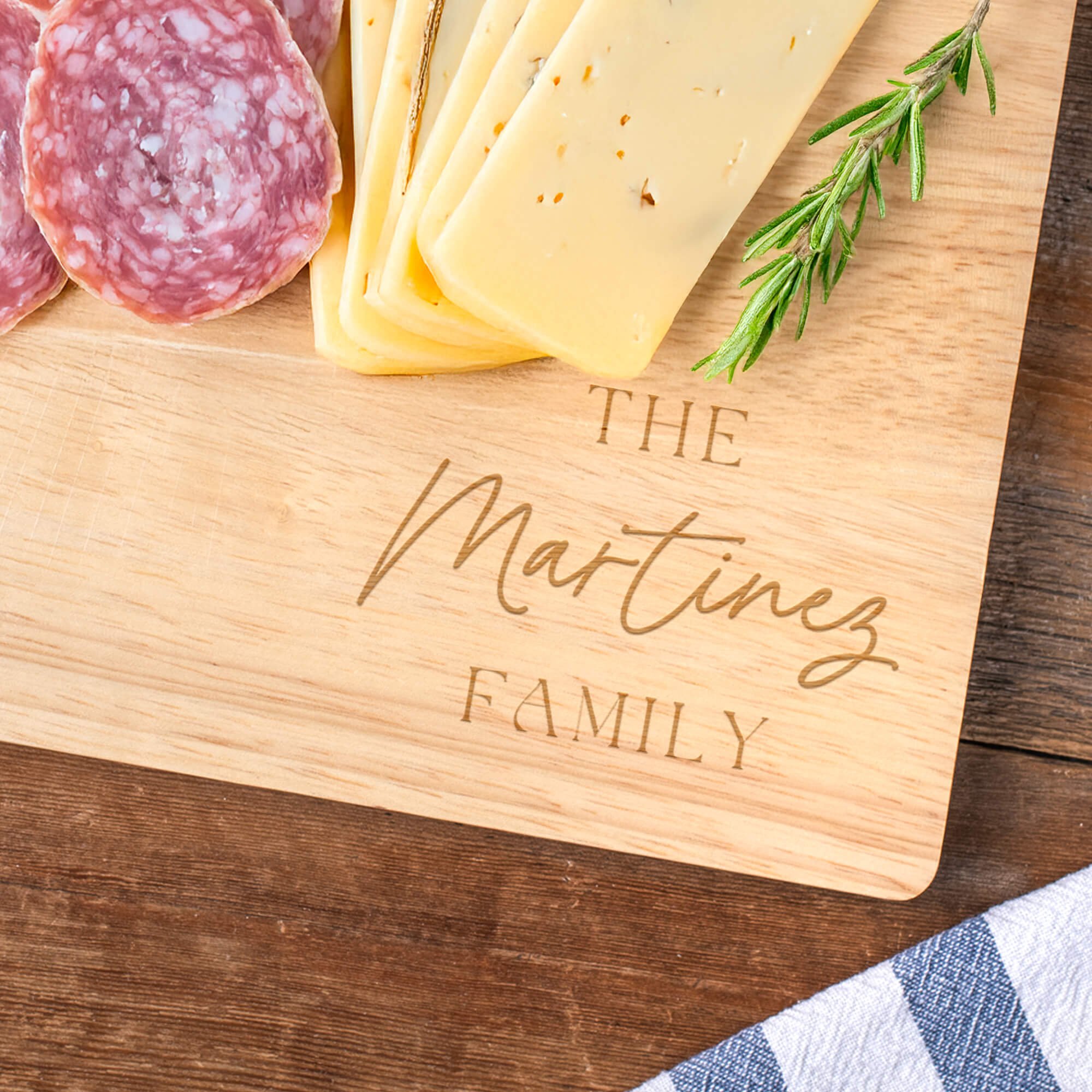 Close-up of engraved maple cutting board personalized with “The Martinez Family,” surrounded by sliced cheese, salami, and a sprig of rosemary on a rustic wooden table.