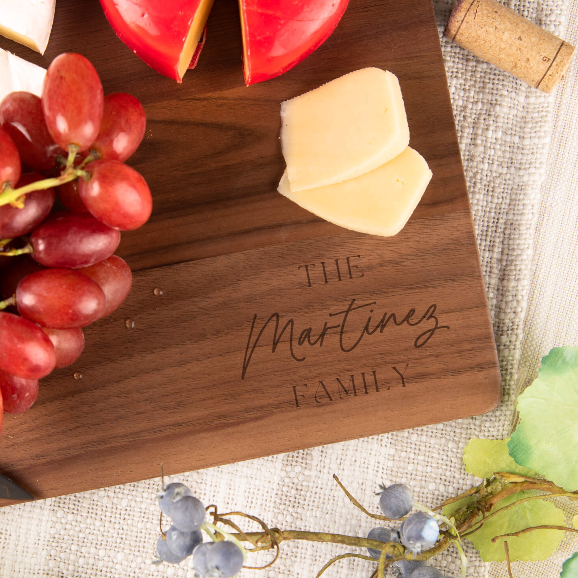 Close-up of a personalized walnut cutting board engraved, surrounded by grapes, cheese, and charcuterie. Custom engraved wedding cutting board or anniversary gift idea.