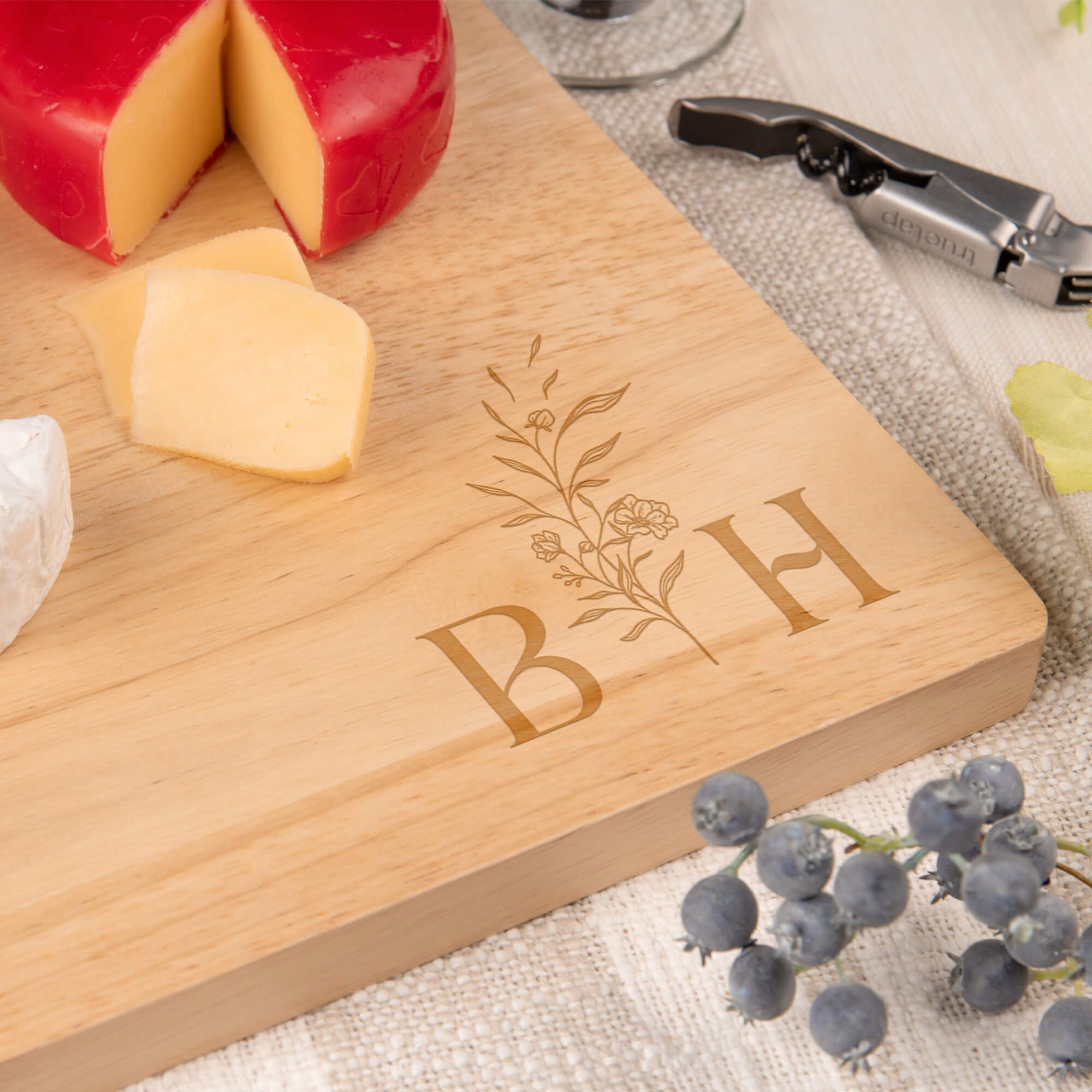 Close-up of a maple cutting board engraved with floral initials “B” and “H,” surrounded by cheese, grapes, and a corkscrew. Elegant custom charcuterie board for weddings or anniversaries.