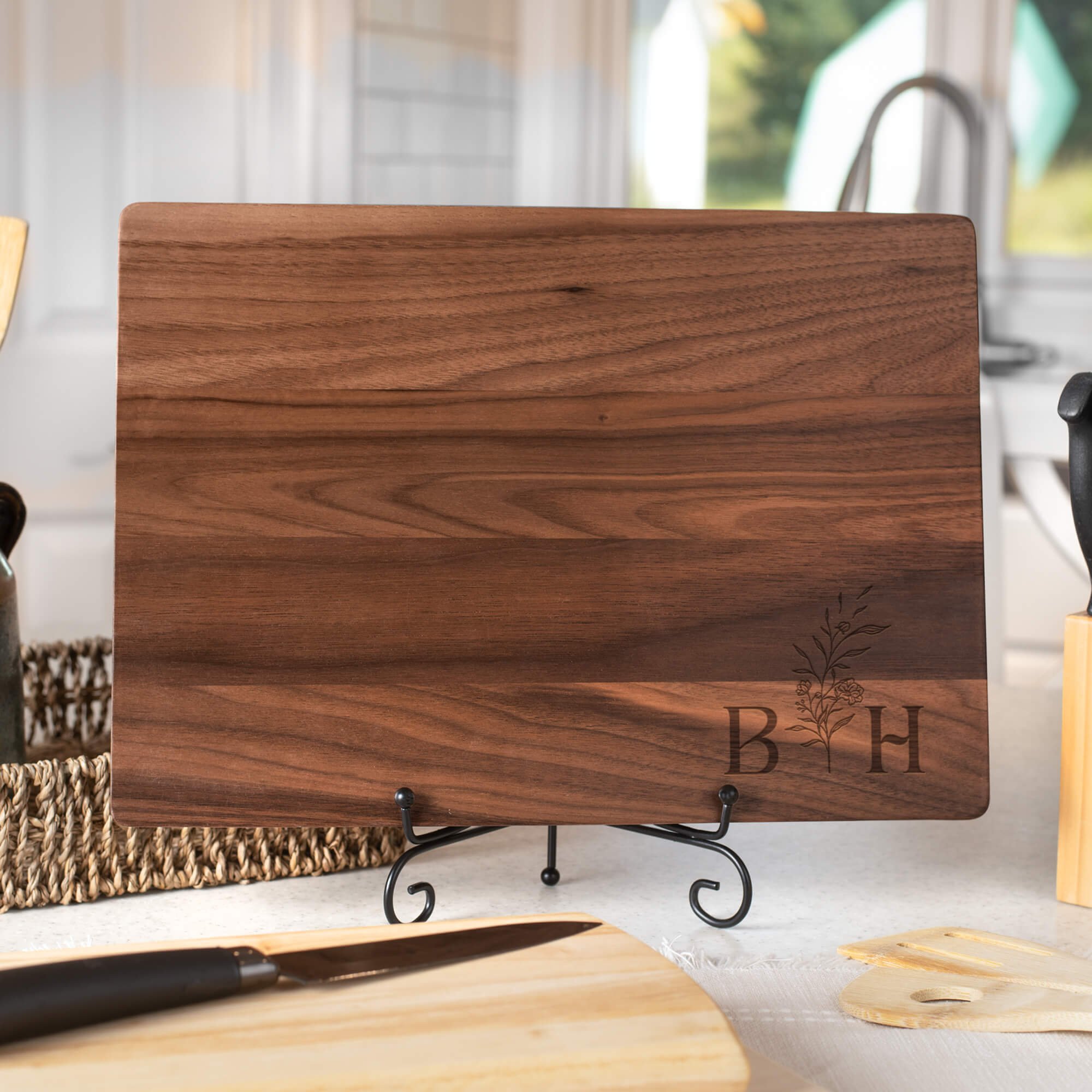 Personalized walnut cutting board engraved with floral initials “B” and “H,” displayed on a black metal stand in a bright kitchen. Custom engraved serving board or monogram wedding gift.