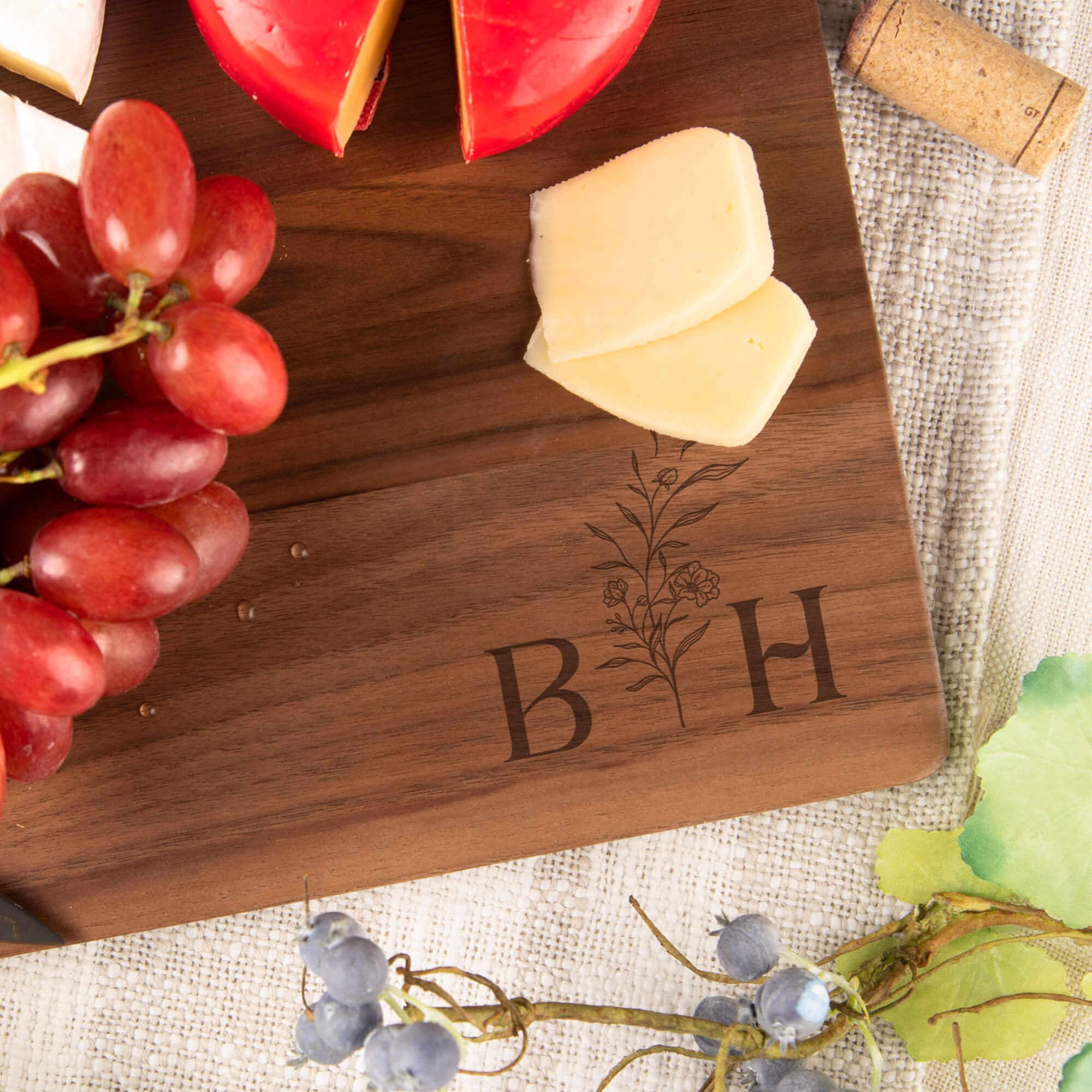 Close-up of engraved walnut cutting board with floral initials “B” and “H,” surrounded by cheese, grapes, and charcuterie. Elegant personalized wood serving board or anniversary gift.