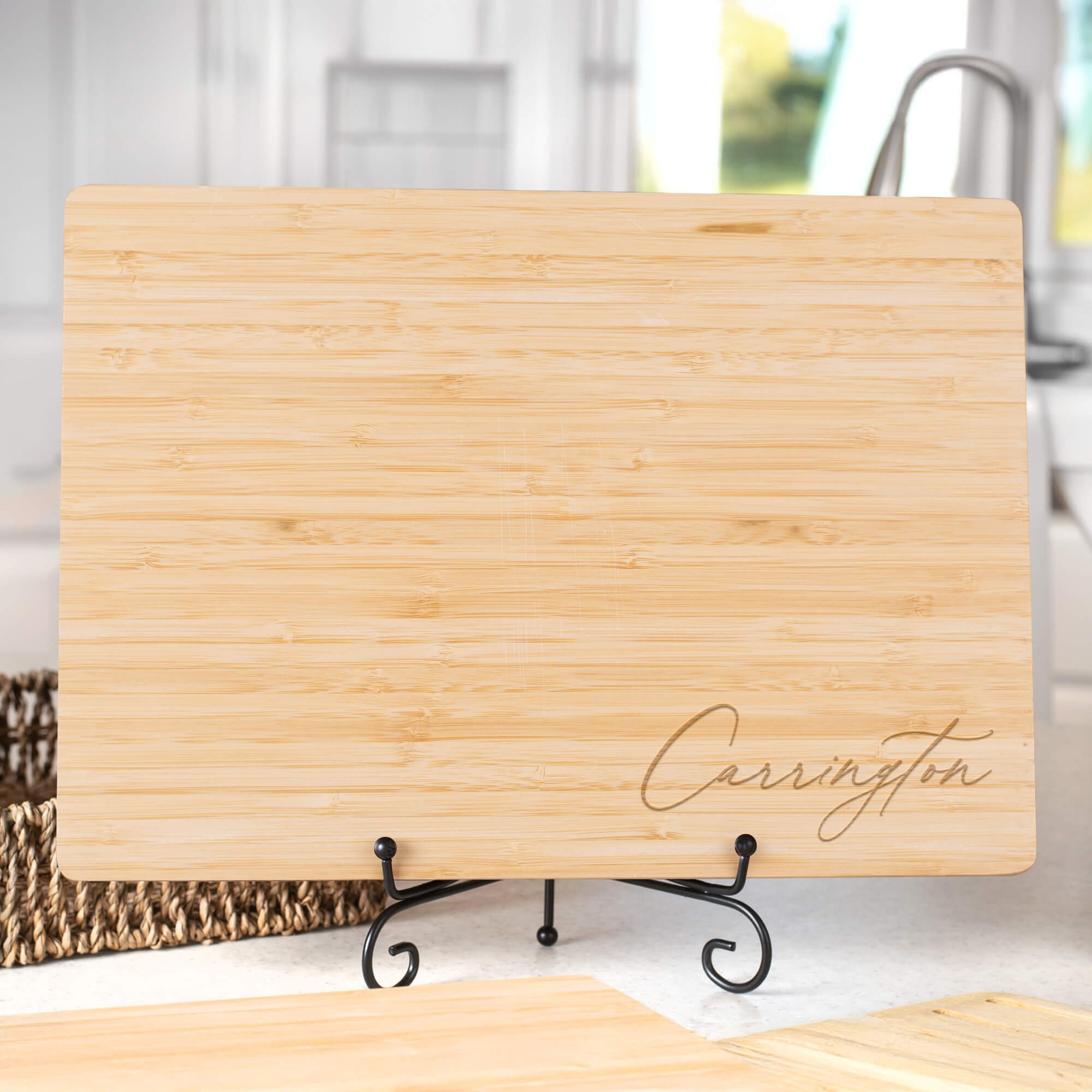 Personalized bamboo cutting board engraved with “Carrington” in elegant script, displayed on a black metal stand in a bright kitchen. Custom engraved serving board or wedding gift.