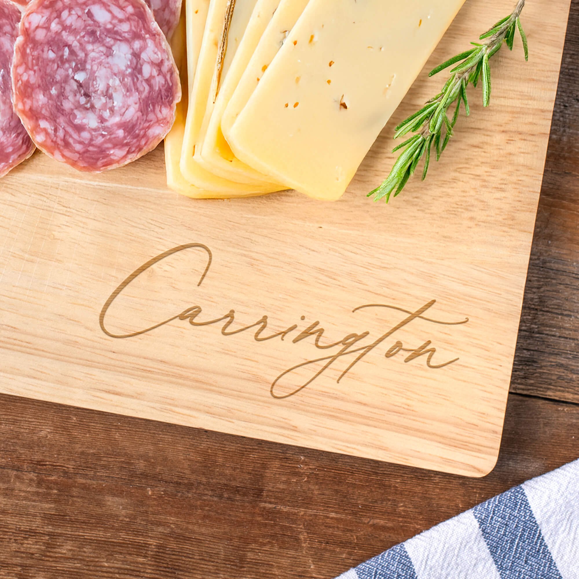 Personalized maple cutting board engraved with “Carrington,” styled with sliced cheese, rosemary, and charcuterie. Custom engraved cutting board for entertaining or gift giving.