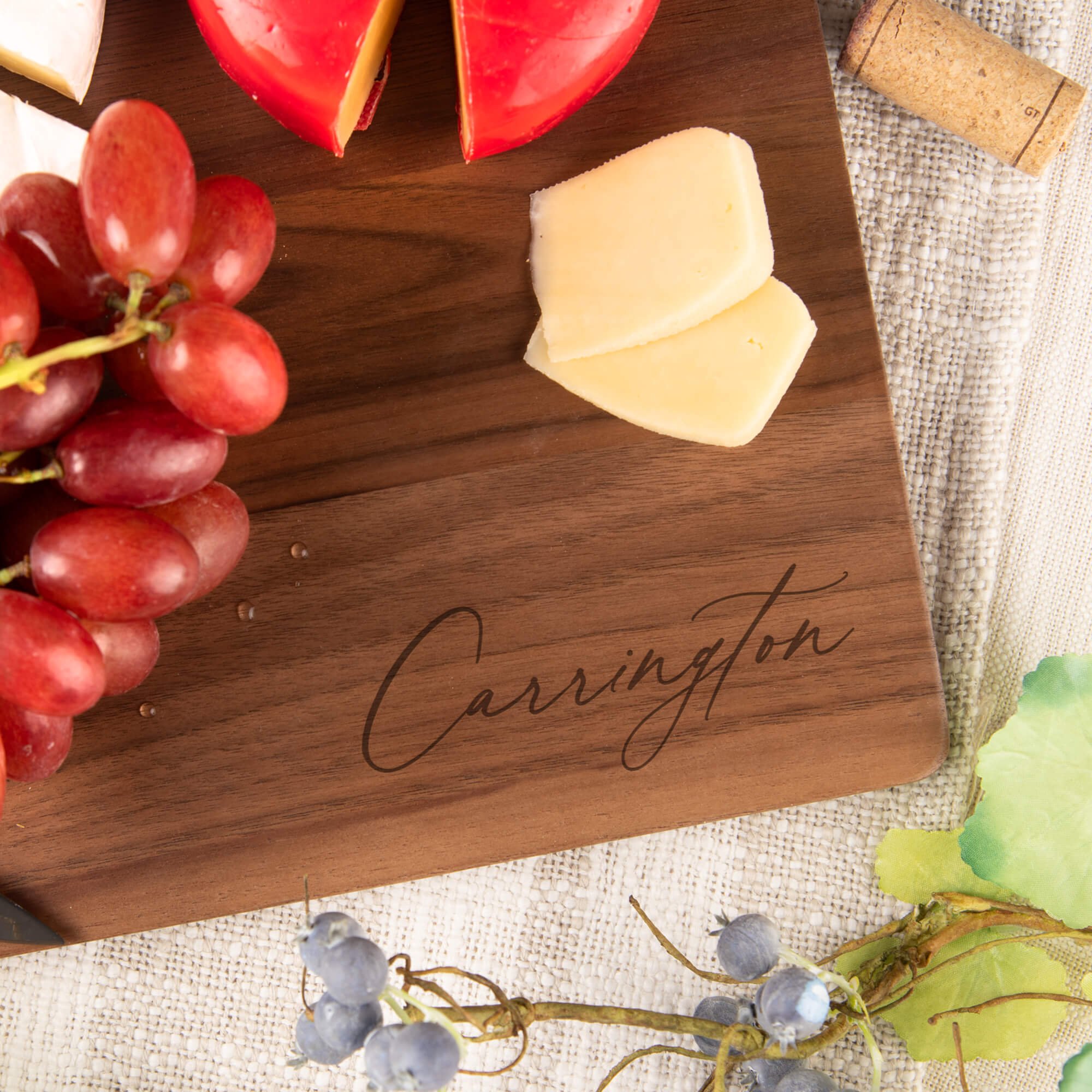 Close-up of personalized walnut cutting board engraved with “Carrington,” styled with cheese, grapes, and wine. Elegant engraved cutting board or housewarming gift for couples.