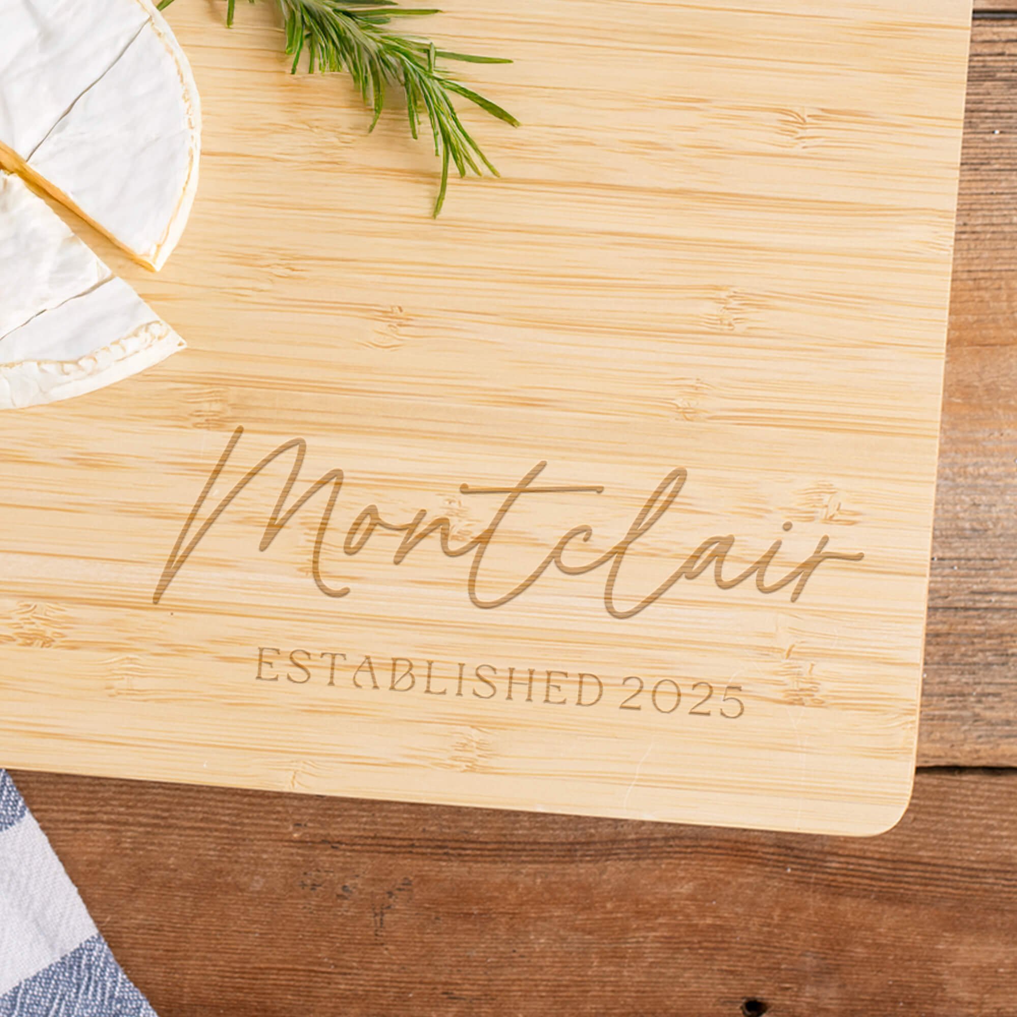 Engraved bamboo cutting board personalized with “Montclair Established 2025,” next to brie cheese and rosemary