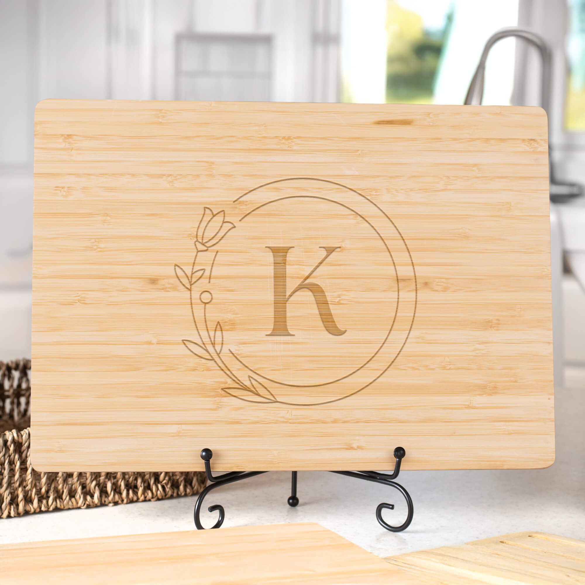 Personalized bamboo cutting board engraved with the letter “K” inside a floral circle design, displayed upright on a black metal stand in a bright kitchen setting.