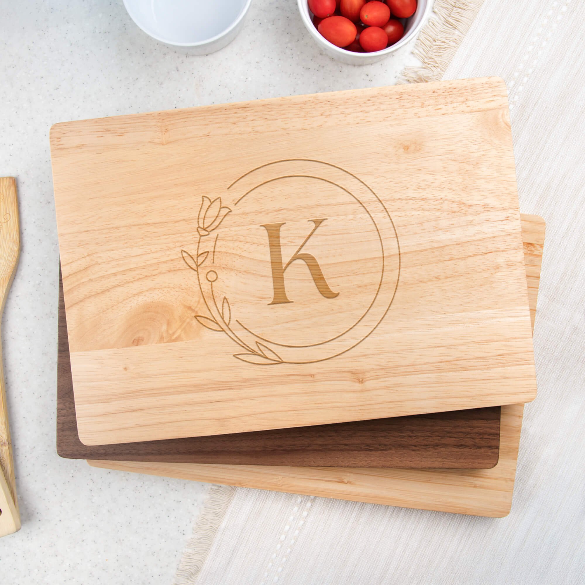 Stack of engraved cutting boards in maple, walnut, and bamboo wood, with the maple board on top featuring the letter “K” inside a floral circle design, displayed on a light kitchen countertop with bowls of cherry tomatoes nearby.