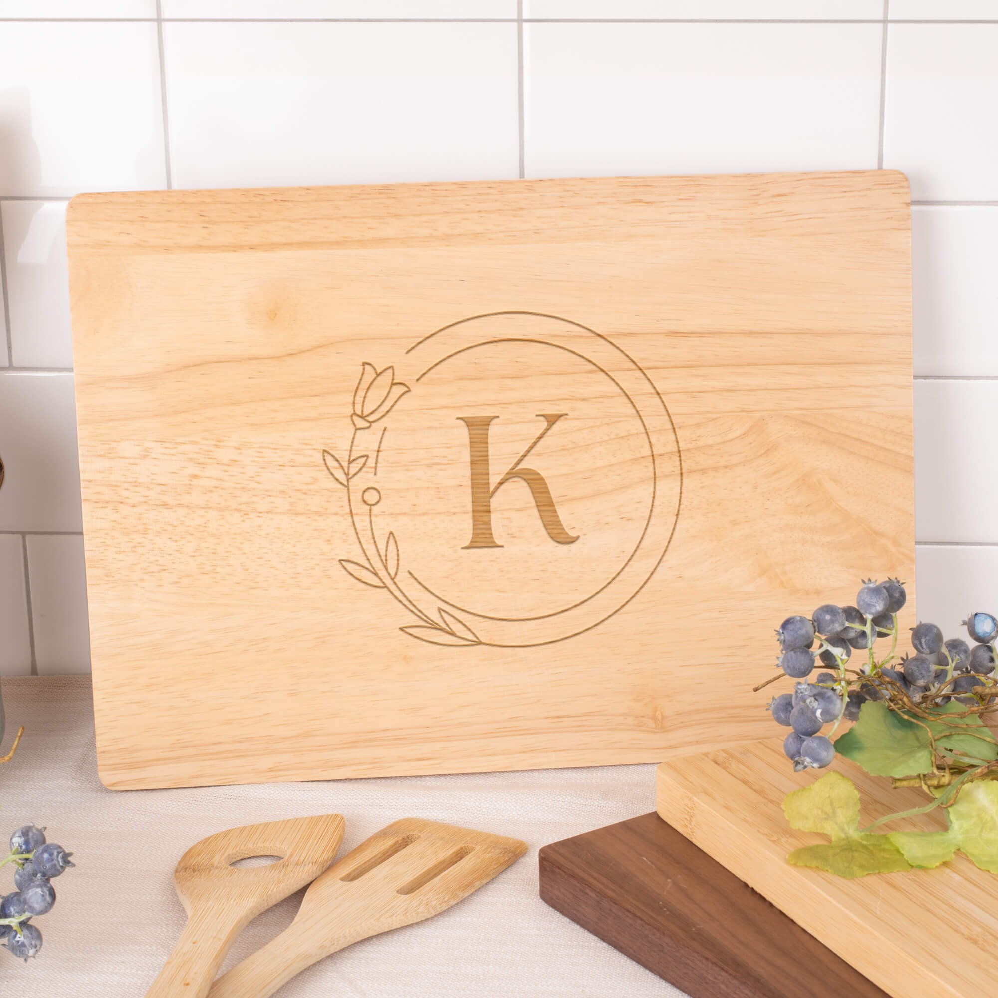 Maple cutting board engraved with the letter “K” inside a floral circle design, displayed against a white tile backsplash with wooden utensils, blueberries, and stacked cutting boards nearby.