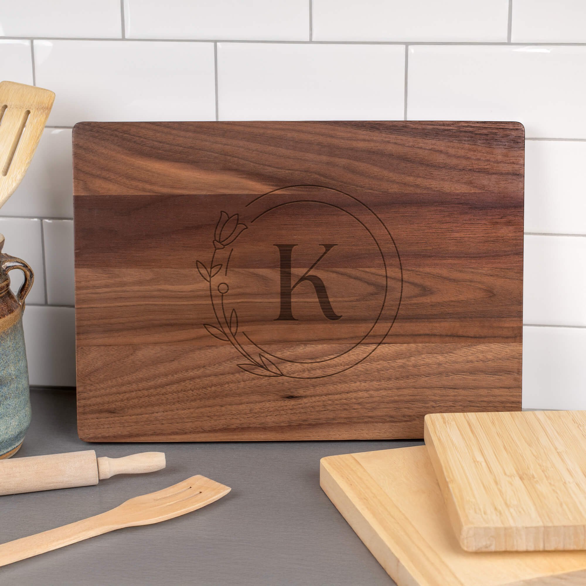 Walnut cutting board engraved with the letter “K” inside a floral circle design, displayed against a white tile backsplash with wooden utensils, a rolling pin, and lighter wood cutting boards nearby.