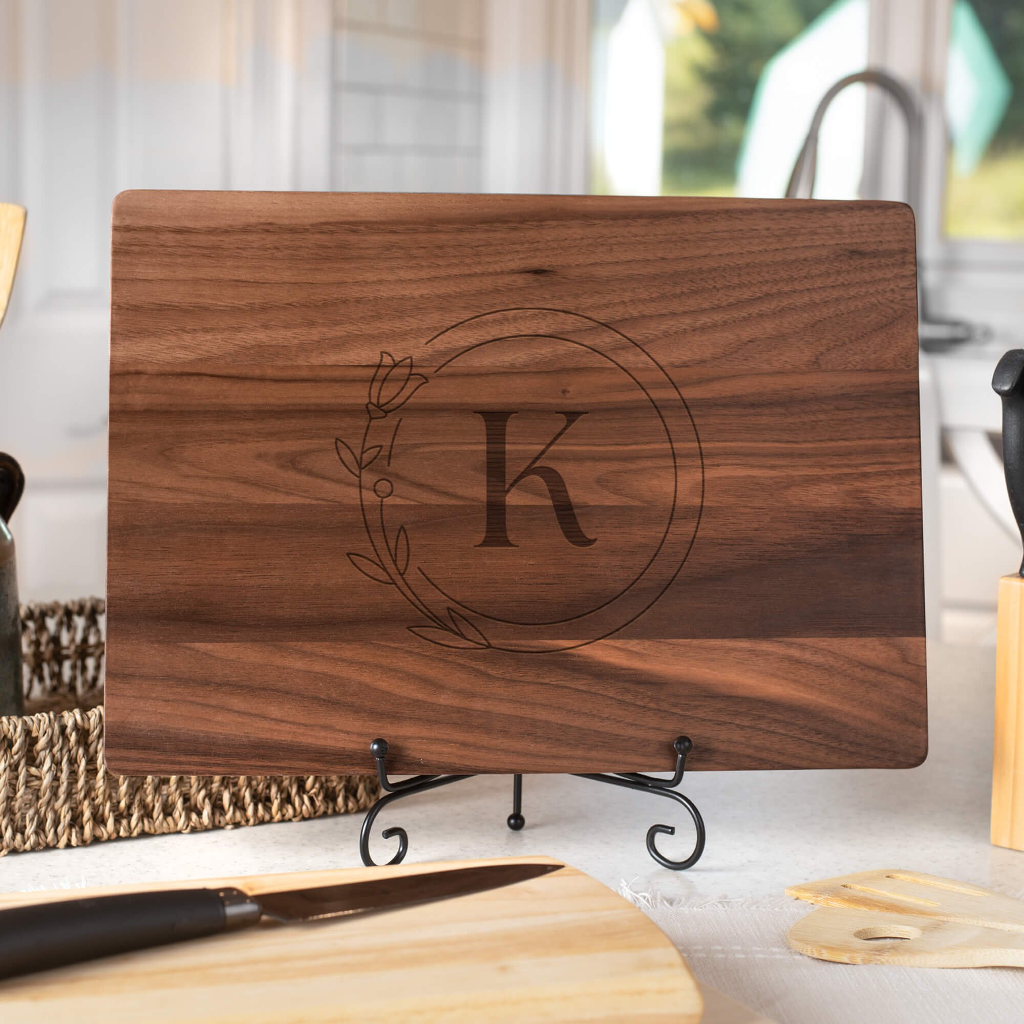 Walnut cutting board engraved with the letter “K” inside a floral circle design, displayed upright on a black metal stand in a bright kitchen with utensils, a knife, and woven basket nearby.