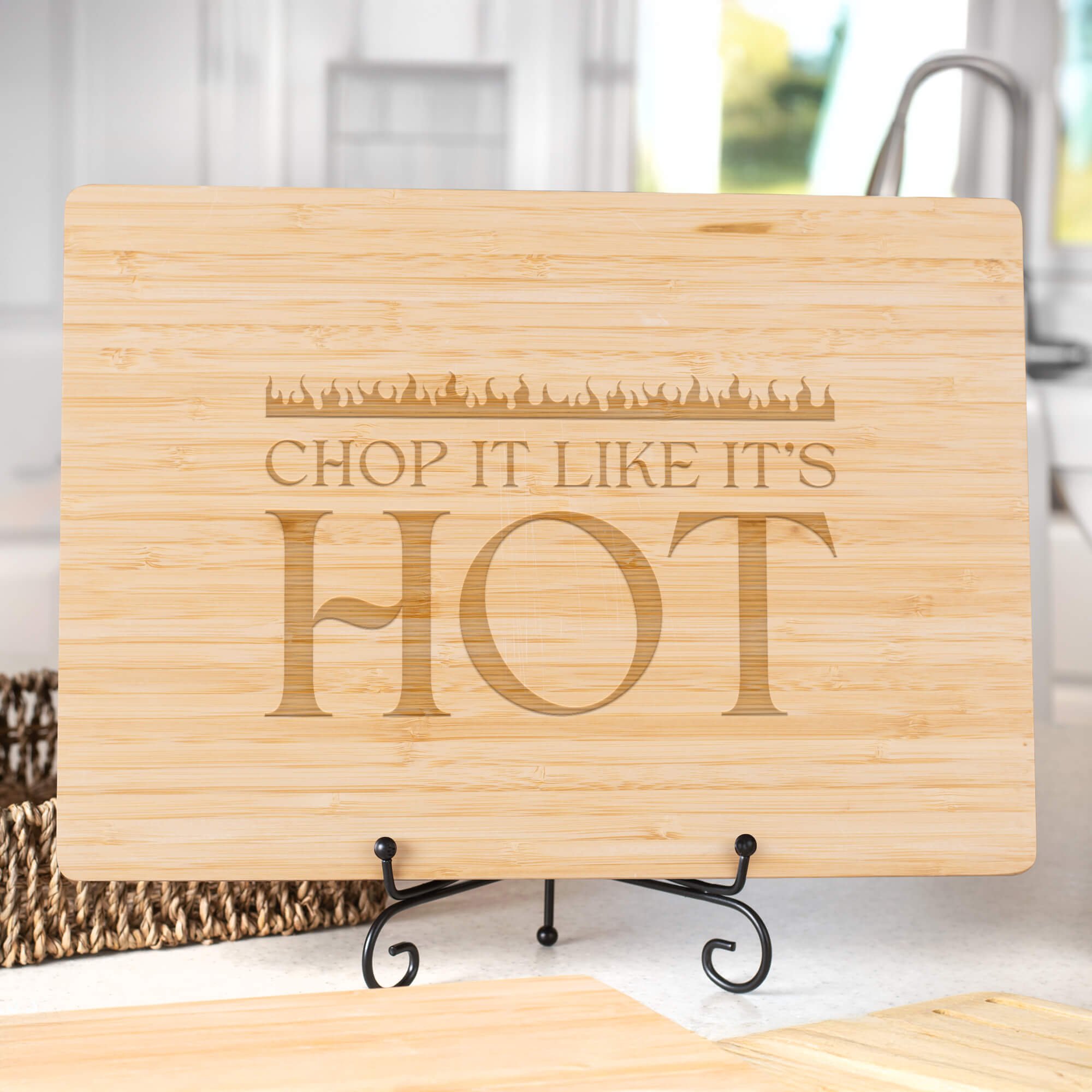 Personalized bamboo cutting board engraved with “Chop It Like It’s Hot,” displayed on a black metal stand in a bright kitchen. Fun cooking gift for home chefs.