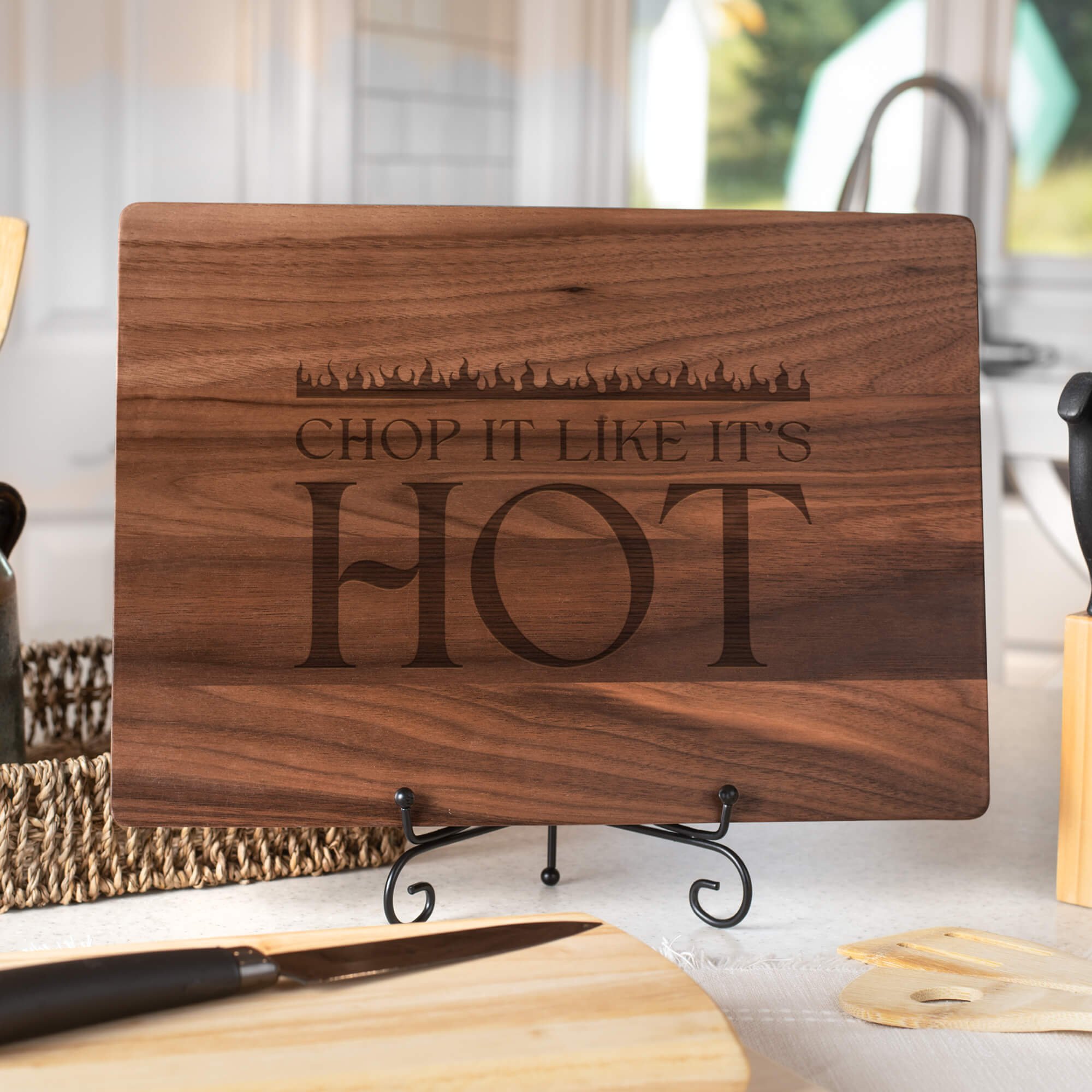 Personalized walnut cutting board engraved with “Chop It Like It’s Hot,” styled with utensils and bamboo boards. Humorous cooking board for BBQ lovers or kitchen décor.