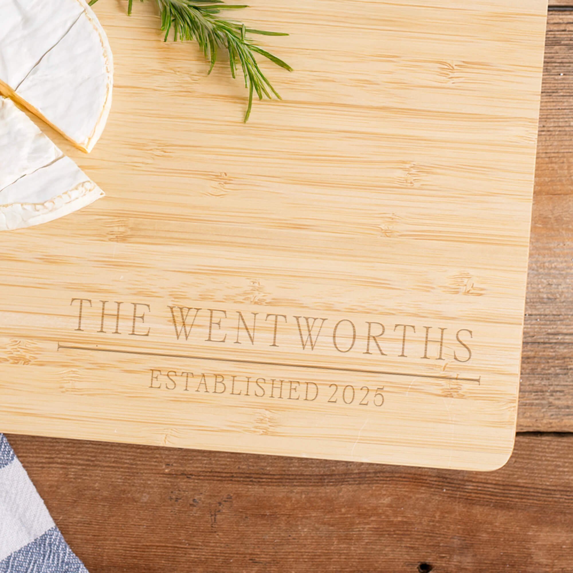 Personalized bamboo cutting board engraved with 'The Wentworths Established 2025' zoomed in to highlight the design next to cheese and rosemary